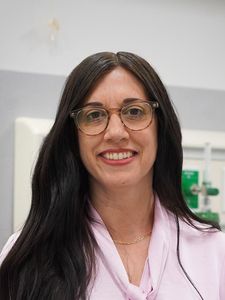A woman with long dark hair and glasses smiles while wearing a light pink blouse and a thin gold necklace.