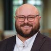 A JCU faculty member, a man with a bald head, beard, and glasses, offers a warm, broad smile for his portrait.