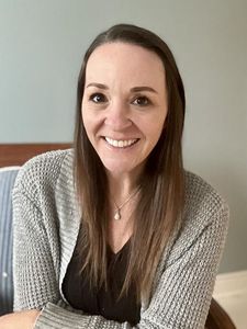 A woman with long brown hair smiles at the camera wearing a gray knit cardigan and a necklace while sitting on a chair.