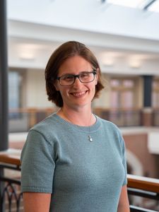 A woman with short brown hair and glasses smiles while standing indoors near a railing with blurred background.
