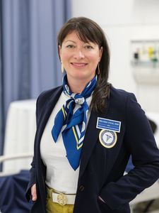 A woman with dark hair wearing a navy blazer, blue scarf, and white shirt stands smiling in a clinical setting.