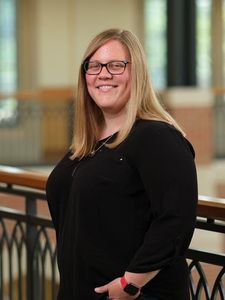 A smiling woman with long blonde hair and glasses stands indoors by a railing, wearing a black top and red watch.