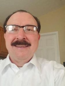 A man with glasses and a mustache wearing a white shirt smiles in a room with a closed white door behind him.