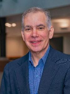 A middle-aged man wearing a black blazer and light blue shirt smiles directly at the camera in an indoor setting.