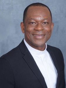 A man with short hair smiles at the camera wearing a striped sweater over a checkered shirt against a plain background.