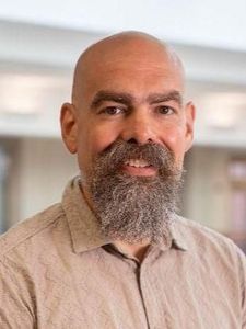 A bald man with a thick gray beard wearing a dark navy polo shirt looks directly at the camera.