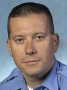 A man with short dark hair wearing a light blue collared shirt looks directly at the camera with a neutral expression.