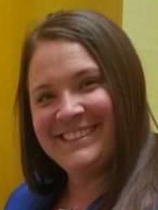 A woman with long brown hair smiles slightly while wearing a blue top against a yellow and beige background.