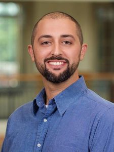 A man with a trimmed beard and short hair smiles while wearing a blue checkered button-up shirt indoors.