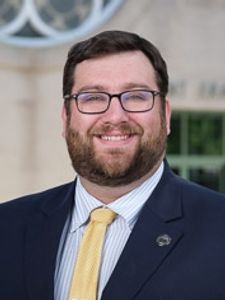 A man with glasses and a beard smiles while wearing a dark suit and yellow tie in front of a building entrance.