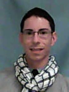 A person with short dark hair and glasses smiles while wearing a white and black patterned scarf and a light-colored top.