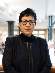 A person with short dark hair and glasses stands indoors wearing a dark jacket over a black turtleneck.