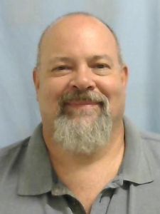 A middle-aged man with a bald head and gray beard smiles slightly while wearing a gray collared shirt against a light blue.