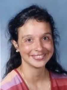 A woman with dark hair pulled back, wearing pearl earrings and a striped top, smiling against a plain blue background.