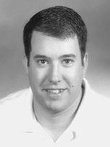 A black and white portrait of a man with short dark hair wearing a light-colored collared shirt, smiling slightly.