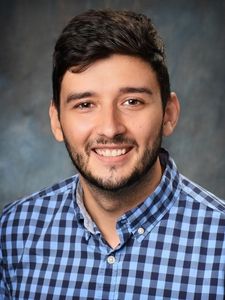A man with dark hair and a beard smiles while wearing a blue and black checkered shirt against a gray background.