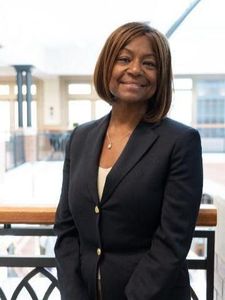 A woman with shoulder-length hair wearing a dark blazer smiles while standing indoors near a railing.