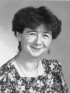 A woman with short curly hair smiles gently, wearing a floral patterned top and dangling earrings against a plain background.