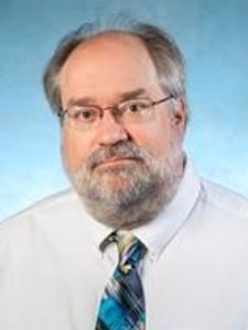 A middle-aged man with glasses, a beard, and mustache wearing a white shirt and a blue patterned tie against a blue.
