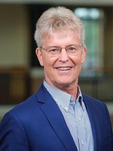 A smiling older man with short gray hair and glasses wears a blue blazer over a striped shirt against a blurred background.