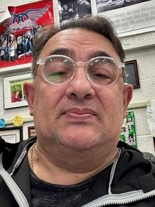 A man wearing clear glasses and a black jacket looks into the camera with a wall of photos and posters behind him.