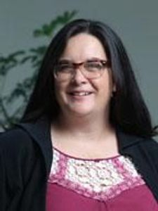 A woman with long dark hair and glasses smiles while wearing a maroon top with a white lace neckline and a black cardigan.