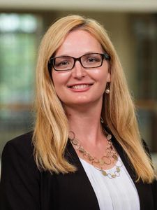 A smiling woman with long blonde hair wearing black glasses, a black blazer, white blouse, and a gold necklace stands.