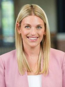 A woman with straight blonde hair wearing a pink blazer and white top smiles in a softly lit indoor setting.