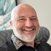 A JCU faculty member, a bald man with a grey beard, beams at the viewer, wearing a black shirt with a patterned collar.