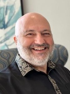A JCU faculty member, a bald man with a grey beard, beams at the viewer, wearing a black shirt with a patterned collar.