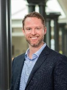 A man with short brown hair and a beard smiles while standing indoors near black columns and a hallway.