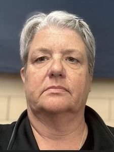 A person with short gray hair and a neutral expression is facing the camera against a background of beige bricks and a dark.