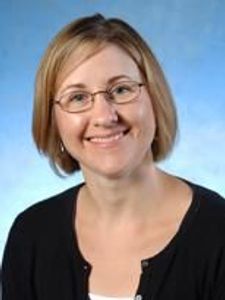 A smiling woman with short blonde hair and glasses wearing a black cardigan over a white top against a blue background.