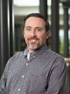 A man with short dark hair and a beard smiles while standing indoors near tall black columns and windows.