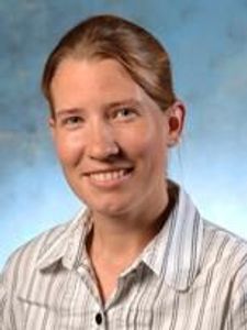A woman with light brown hair pulled back is smiling and wearing a white shirt with thin gray stripes against a blue-gray.