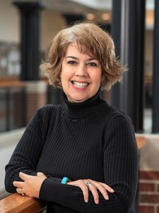 A woman with short light brown hair smiles with her arms crossed resting on a wooden railing indoors.