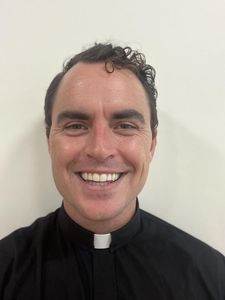 A man with curly dark hair smiles at the camera wearing a black clerical shirt with a white collar tab.