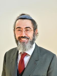 A man with a gray beard and glasses smiles while wearing a gray suit, white shirt, red tie, and a patterned kippah.
