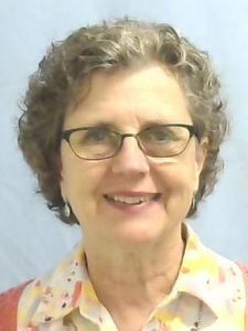 A woman with short curly hair and glasses smiles wearing a patterned blouse and a coral cardigan against a plain background.