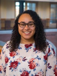 A smiling woman with curly dark hair, glasses, a patterned top, and a green cardigan stands against a plain wall.