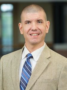 A man with short hair wearing a beige suit jacket, white shirt, and blue striped tie smiles in an indoor setting.
