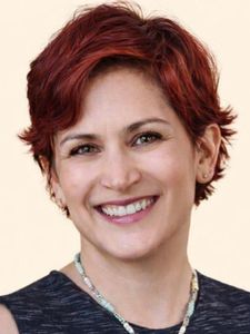 A person with short red hair smiles warmly, wearing a dark top and a beaded necklace against a light background.