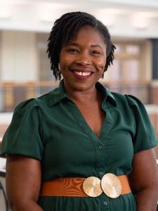 A woman with braided hair wearing a brown sweater and a large white beaded necklace stands indoors in front of a white wall.