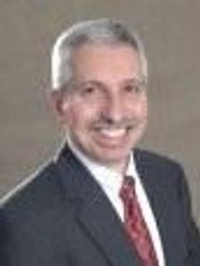 A man with short gray hair and a mustache wearing a dark suit, white shirt, and red tie smiling against a plain background.