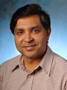 A man with dark hair and brown skin wearing a checked shirt smiles gently against a blue gradient background.