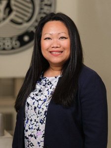 A JCU faculty member with dark hair smiles warmly for her portrait, wearing a navy blazer and white floral blouse.