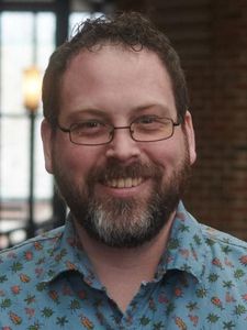 A man with dark curly hair, glasses, and a beard smiles while wearing a maroon collared shirt against a gray background.
