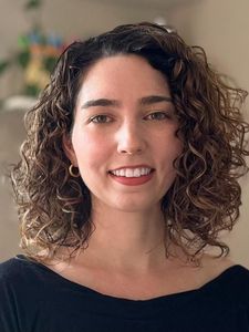 A woman with curly shoulder-length hair smiles softly wearing a black top and gold hoop earrings against a blurred.