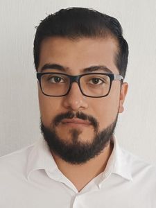 A man with dark hair, beard, and glasses wearing a white collared shirt stands against a plain white wall.
