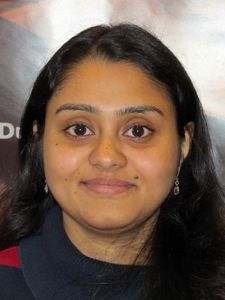 A woman with long dark hair and earrings smiles slightly while sitting in front of a poster with text and images.